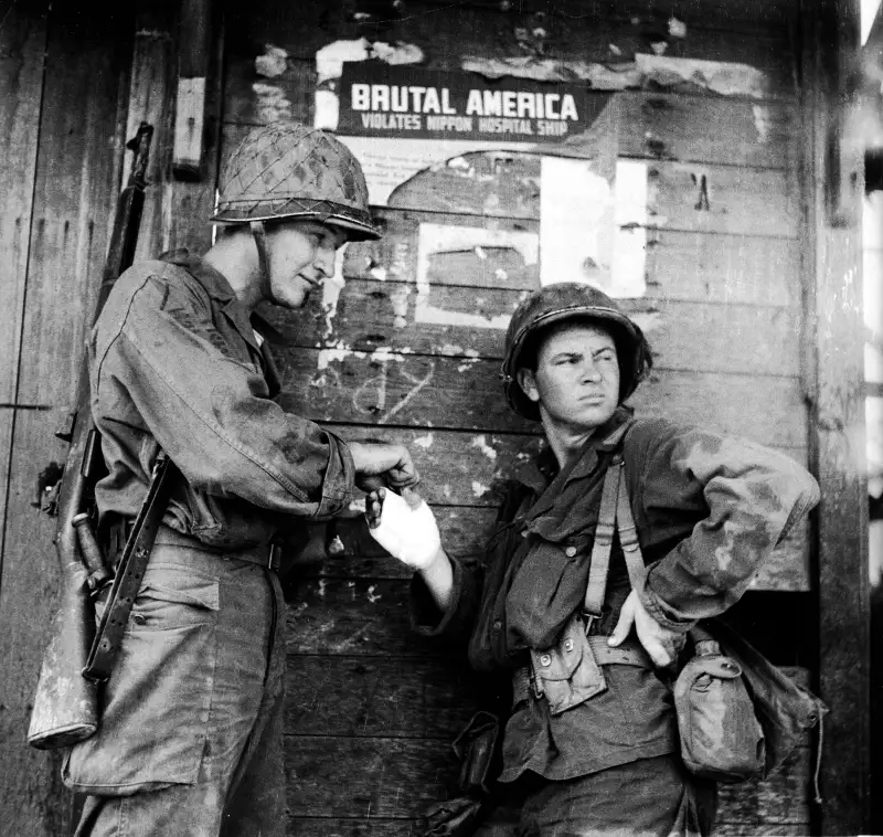 Field Dressing
An American soldier stands nonchalantly while another bandages his hand during the campaign to retake the Philippines from occupying Japanese forces, Leyte, Philippines, October 1944. The pair lean against a wall which has a torn Japanese propaganda poster which reads, Brutal America Violates Nippon Hospital Ship.