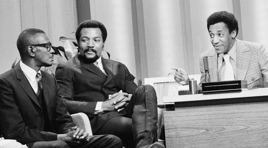 Bill Cosby, filling in as guest host on  The Tonight Show,  interviews Satchel Paige as football legend Jim Brown looks on, amused.