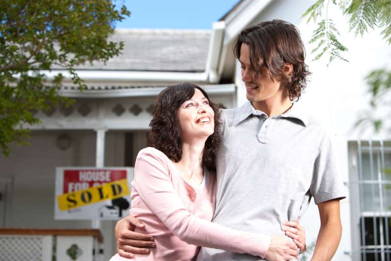 Man and woman embracing in front of house with sold sign