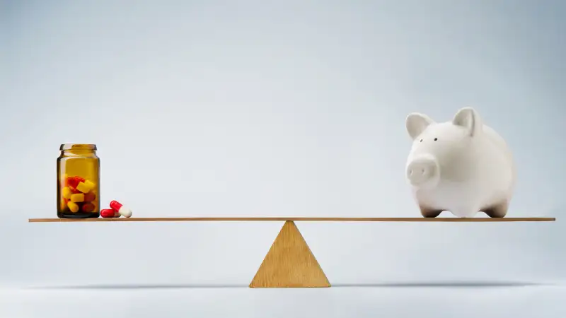 piggy bank and bottle of pills balancing on either side of a seesaw