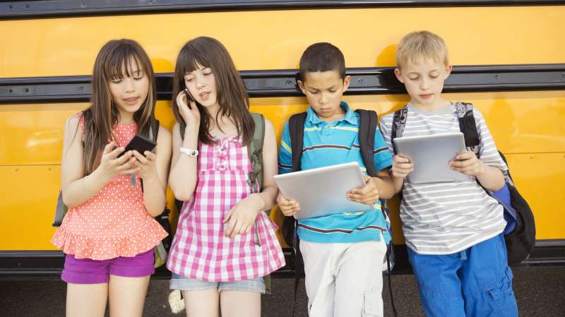 School kids using smartphones and tablets in front of school bus
