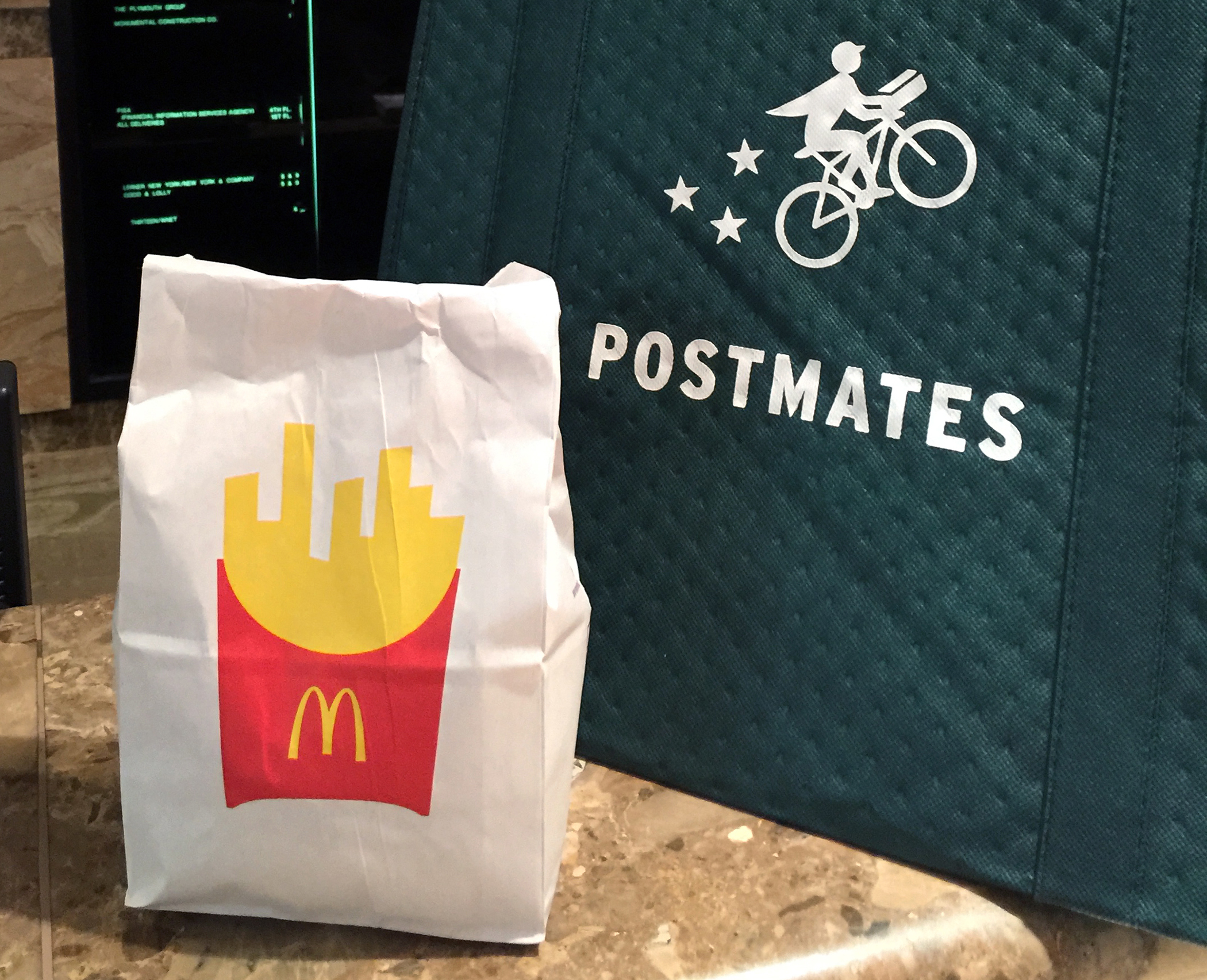 A bag of food from McDonald's ordered through the Postmates service next to a Postmates delivery bag.