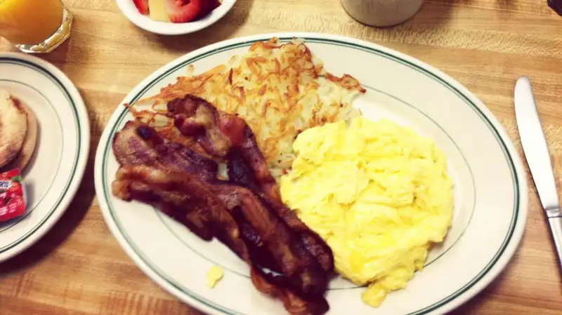 Bacon on plate with scrambled eggs and hash browns, with coffee and fruit salad and orange juice