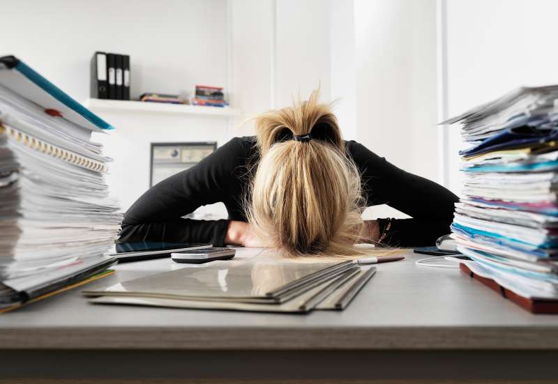 blond woman in office with head on desk and stacks of files