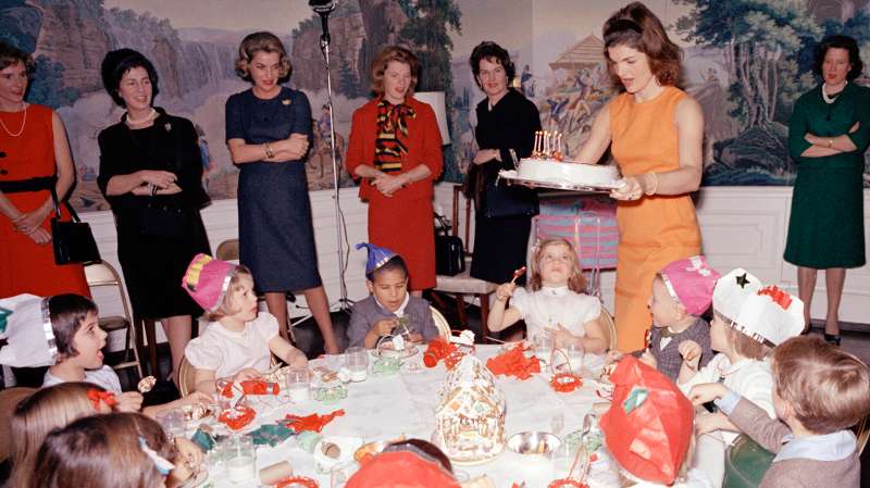 First Lady Jacqueline Kennedy carries Caroline Kennedy’s cake to the table during a joint birthday party for Caroline and John F. Kennedy, Jr., in the President’s Dining Room (Residence), White House, Washington, D.C., November 27, 1962. Avery Hatcher, son of Associate Press Secretary, Andrew T. Hatcher, sits left of Caroline; Maria Shriver (with back to camera) sits at left in foreground. Also pictured: Janet Auchincloss; White House butler, John W. Ficklin.