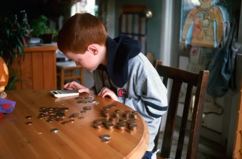 7 year old boy counting his money using a calculator at home in kitchen, UK