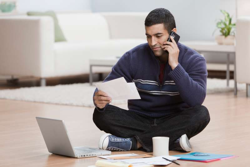 Hispanic man sitting on floor using laptop in living room and looking at mail