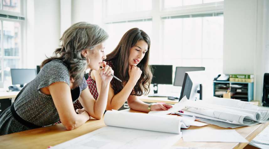 Two businesswomen leaning on table discussing architectural plans in office