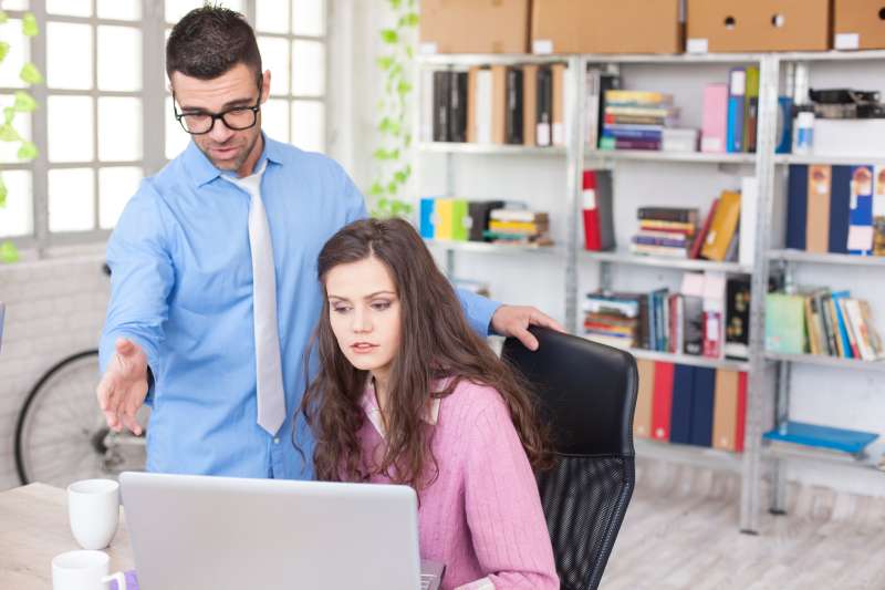 Young female assistant having problems at workplace. Young woman sitting on desk and working on laptop. The manager showing her some errors on files. Man with eyeglasses, blue shirt and white tie. As backgound windows with plants, shelves with boxes and folders, and part of a bike. Cups on desk.