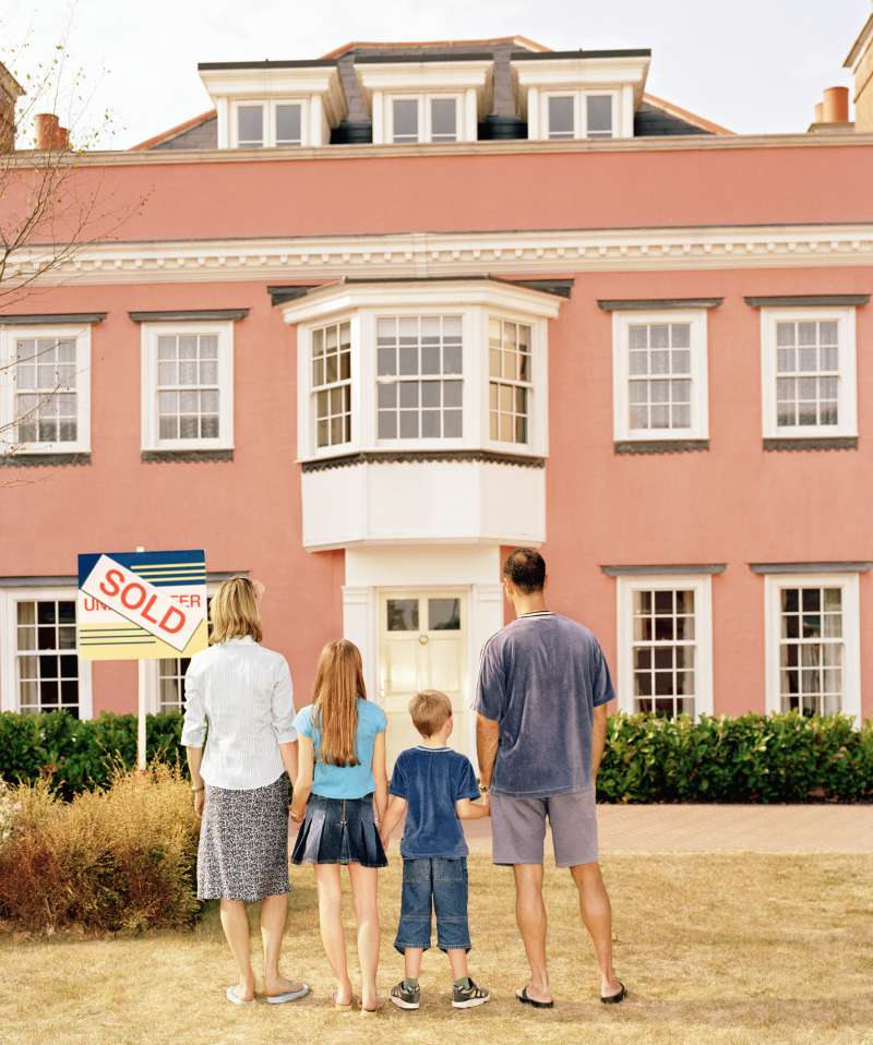 Family holding hands in front of house with 'sold' sign, rear view