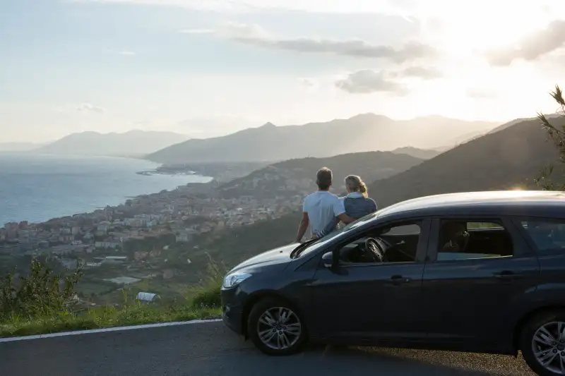 Couple relax against car, look past village to sea