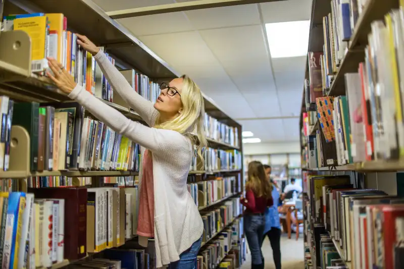 Young female librarian arranging books on library shelves