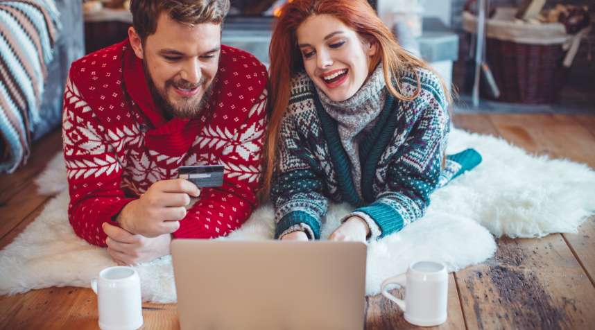 Couple on vacation at mountain cabin. Sitting on the floor on a blanket by a fireplace in a cozy  living room on Christmas. Using laptop to buy gifts for friend and family. Wearing festive knitted sweaters. Austrian Alps.