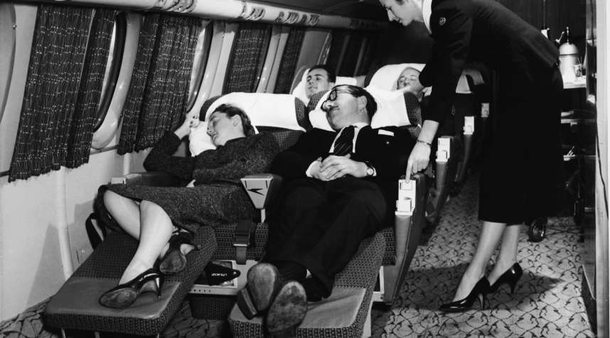 Interior view of the first class compartment of a commercial passenger plane shows a flight attendant as she bends forward to adjust the seat of a sleeping man, 1950s. A woman sleeps beside him, as do the passengers behind him. (Photo by /Getty Images)