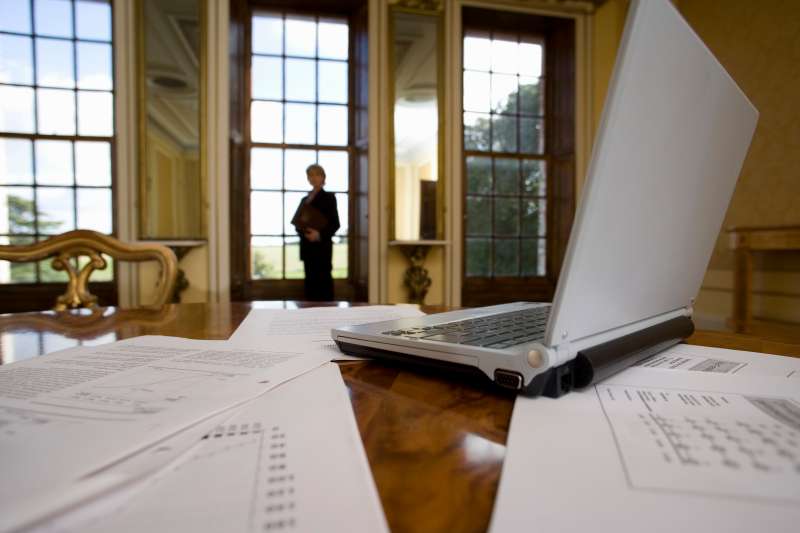 Laptop computer and paperwork on table, woman by window in background, low angle view