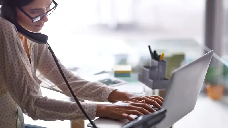 Young woman talking on the phone as she types on her computer.