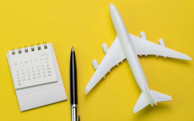Flat lay of travel and tourism concept, small white clean paper calendar with black pen and toy airplane on vibrant yellow background
