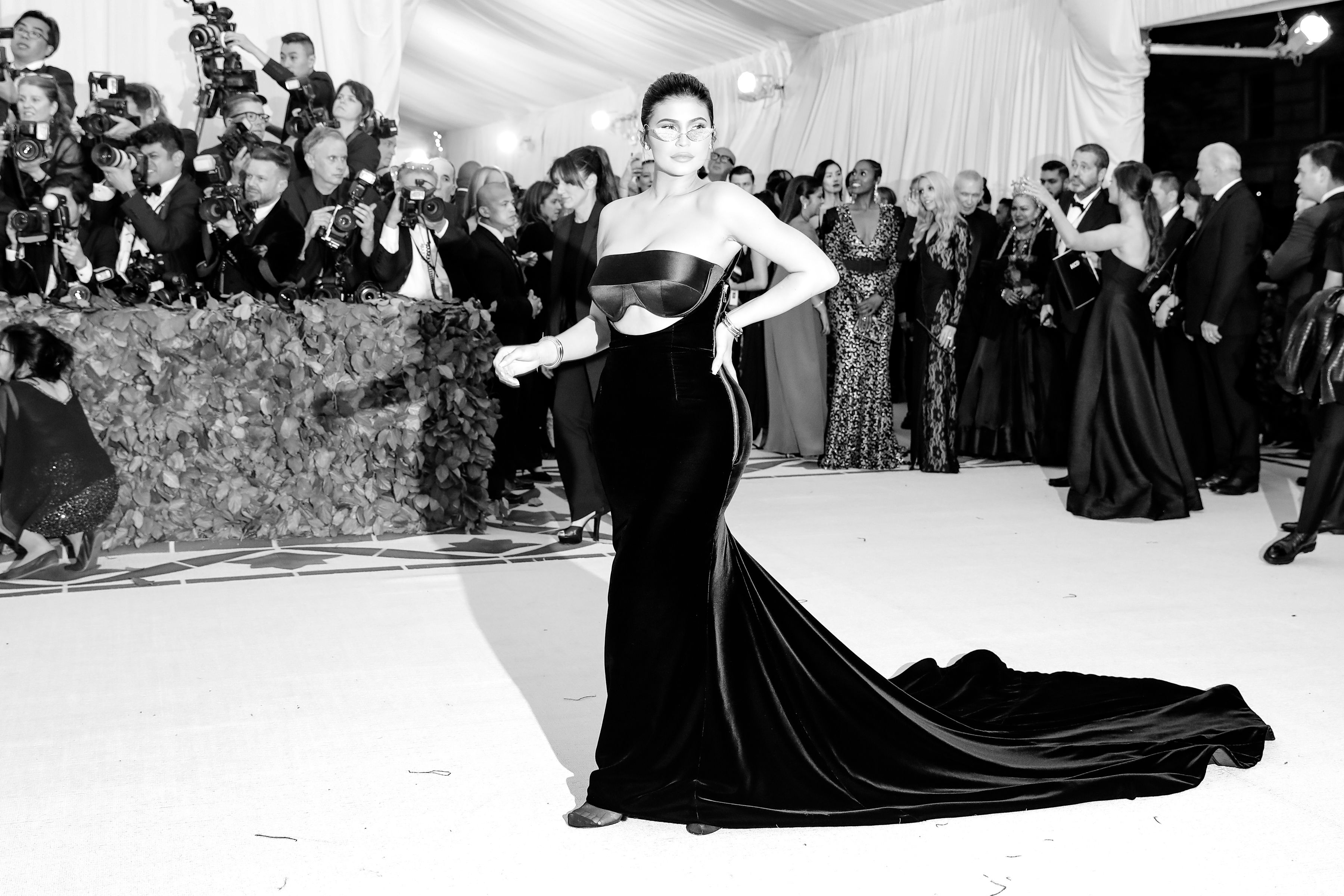 A black and white photo of Kylie Jenner on the red carpet, wearing a floor-length black gown with a long train.