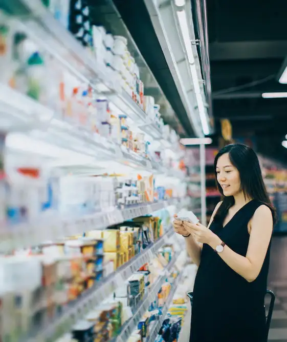 Asian pregnant woman grocery shopping in supermarket and reading nutrition label on a packet of cheese