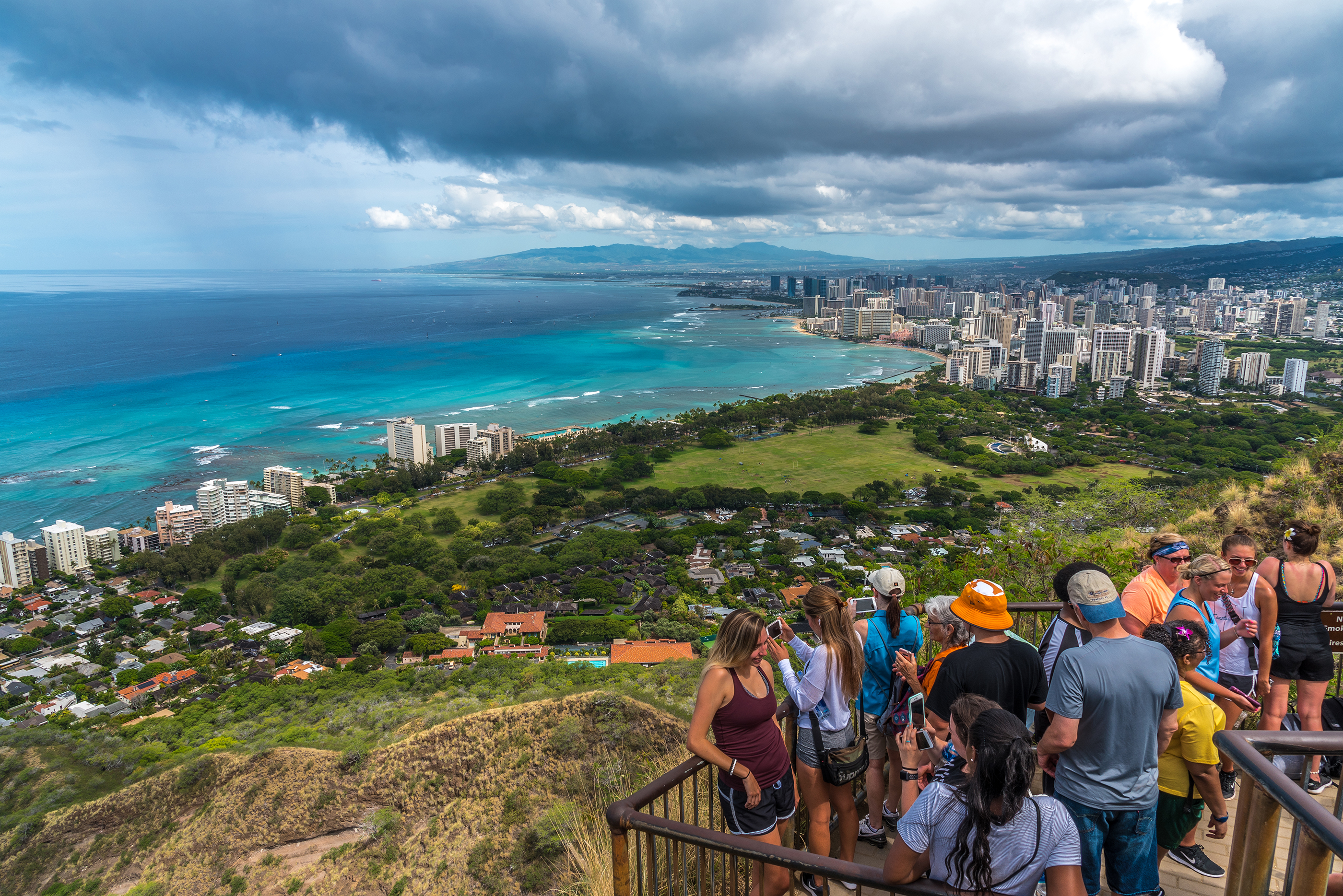 Agroup of tourists take selfies and photos of Waikiki and greater Honolulu from the top of the Diamond Head crater landmark.