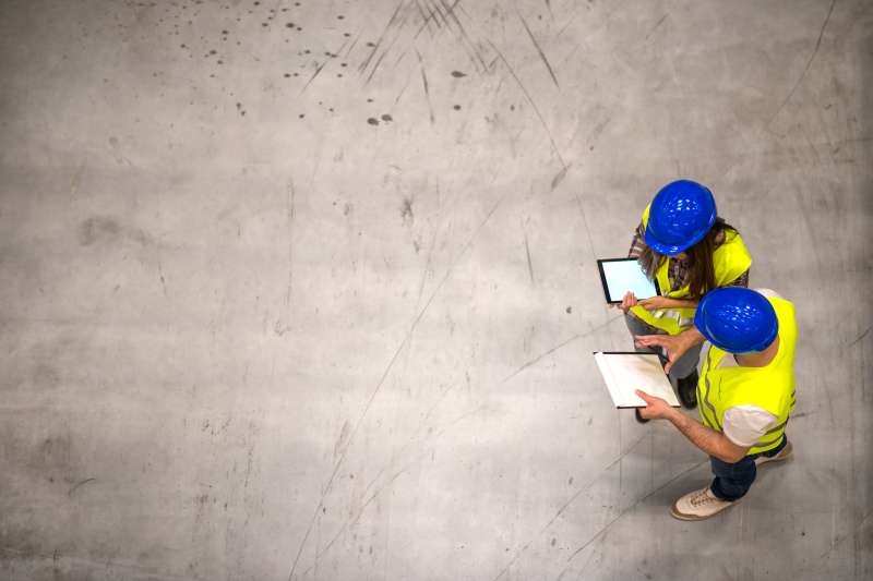 Top view of two construction workers wearing hardhats and reflective jackets holding tablet and checklist on gray concrete background. Group of engineers sharing ideas about the project.