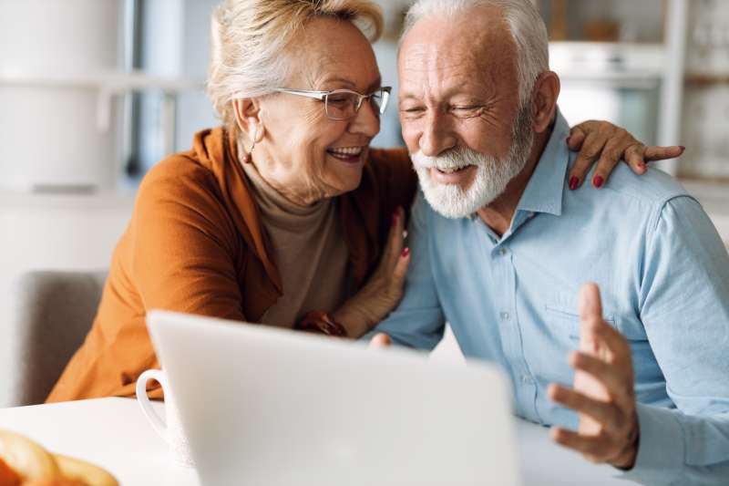 A man and a woman of retirement age embracing and celebrating. He is looking at a computer and she is looking at him.
