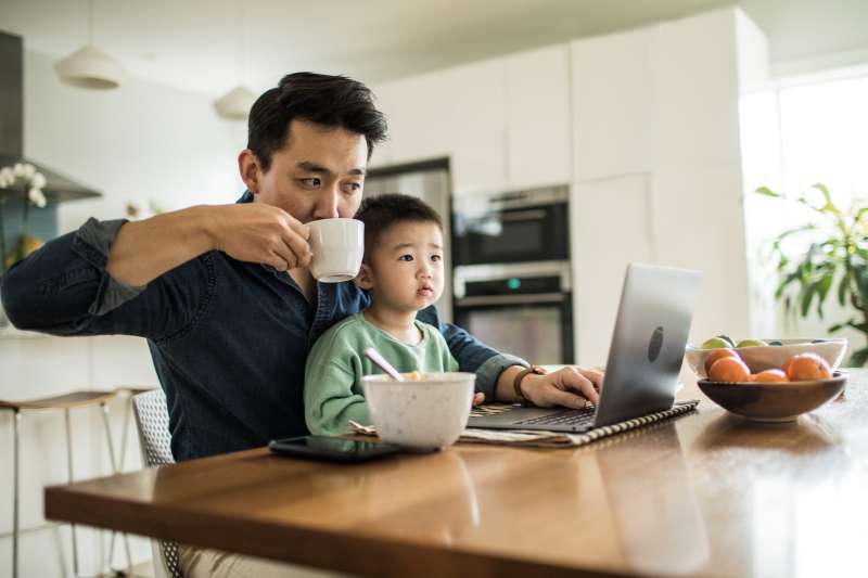 Father with young son on his lap while he looks at his computer at the kitchen table