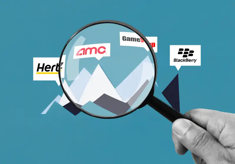 A hand holding a magnifying glass inspecting AMC, Hertz, GameStop and BlackBerry Stocks