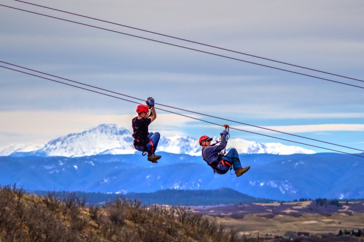 Two people zip-line with a beautiful view of Castle Rock Colorado in the background