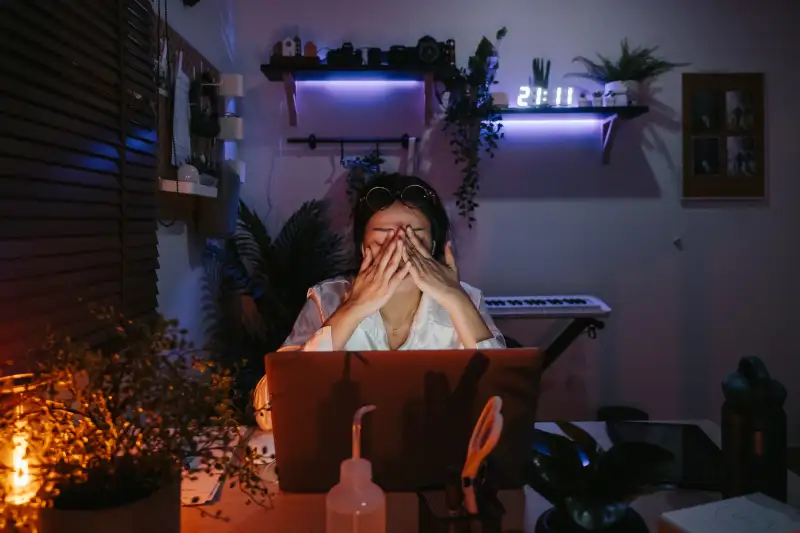 Woman closes her eyes while working late on her computer in a dark room with a clock that reads 21:11 hours.