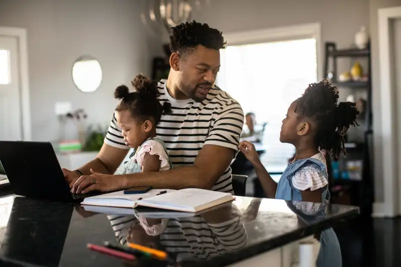 Father multitasking working from home while holding toddler and talking to his older daughter