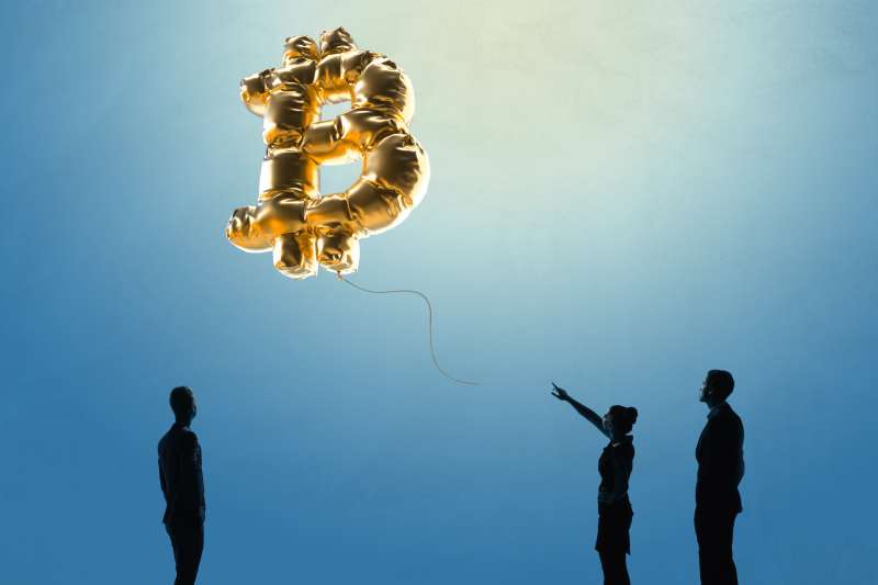 Three people watch from below as a bitcoin balloon rises in the sky