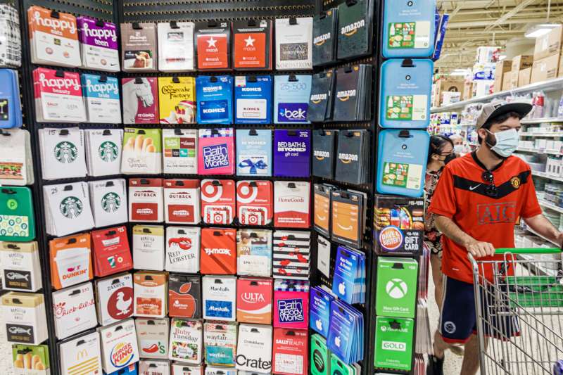 A stand with many gift cards inside a department store. A man is at the side with a shopping cart.