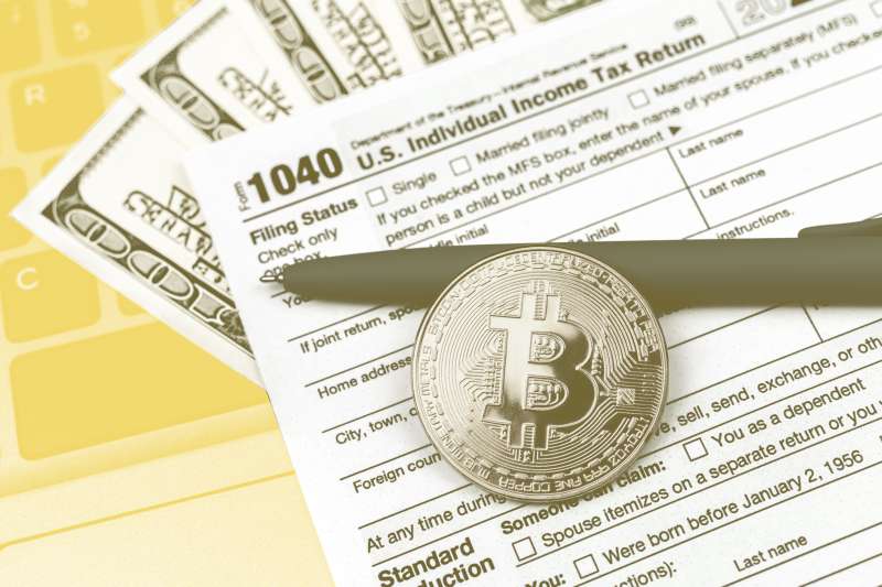 One bitcoin laying on top of a 1040 Tax Return form with dollar bills and a calculator in the background.