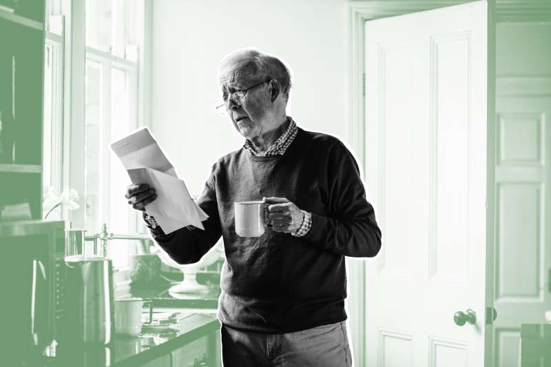 Senior man is standing in the kitchen of his home with bills in one hand and a cup of tea in the other