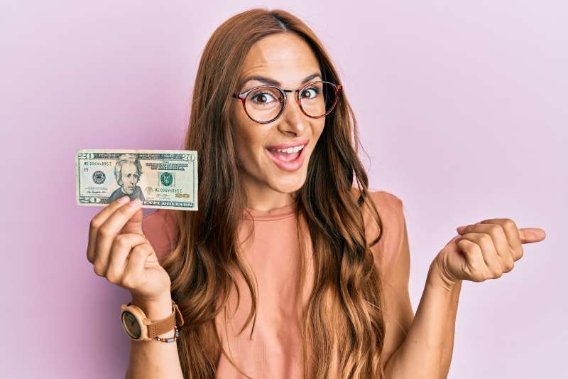 Young brunette woman holding 20 dollars banknote pointing thumb up to the side smiling happy with open mouth