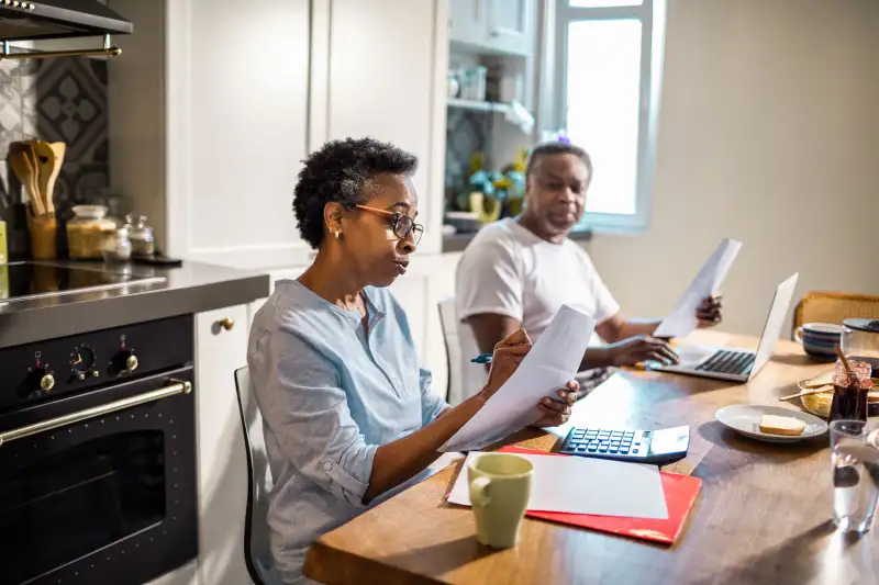 Older Couple Sitting At Kitchen Table Looking At Documents