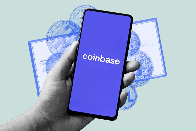 A hand holding a smartphone with the coinbase app open, and a paycheck and multiple crypto currency coins in the background
