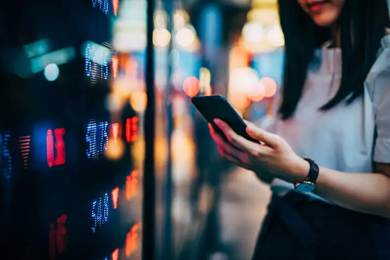 Person Looking At Smartphone In Front Of Screen With Stock Market Graphics