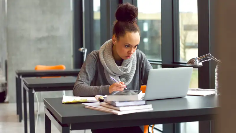 Young student taking notes from laptop and books for her study in library.