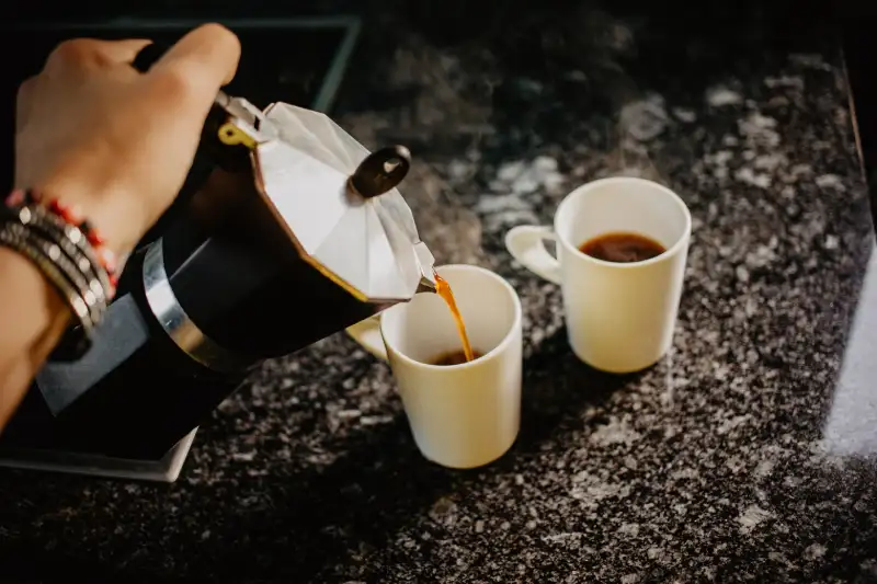 Preparing fresh coffee in moka pot on electric stove. Pouring hot steaming coffee into two white mugs.