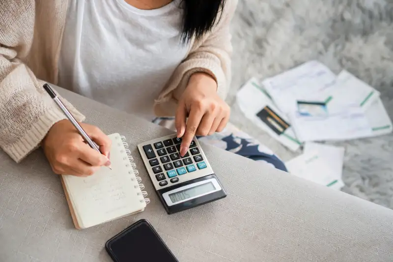 Woman writing a list of debt on notebook calculating her expenses with calculator