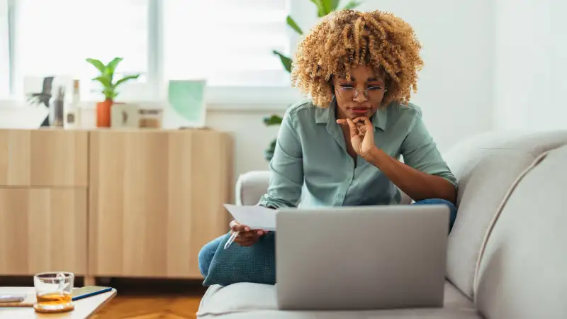 Woman sitting on a couch looking over a piece of paper and working on her laptop