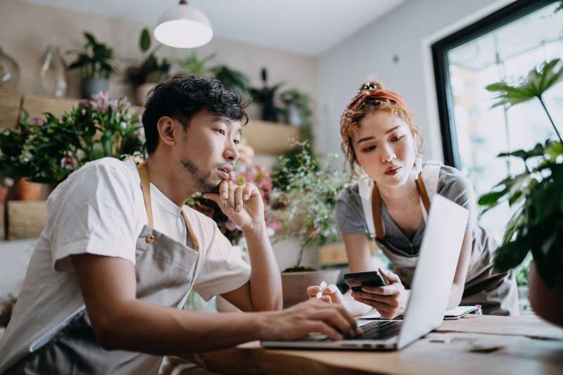 Concentrated young Asian couple, the owners of small business flower shop, discussing over laptop on counter against flowers and plants. Start-up business, business partnership and teamwork. Working together for successful business