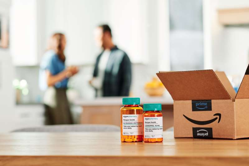 An open amazon delivery box with two prescription pill bottles from Amazon Pharmacy on a table, with a couple in the background