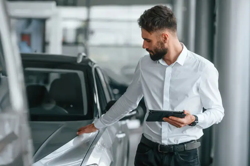 Touching the surface of automobile. Young man in white clothes is in the car dealership