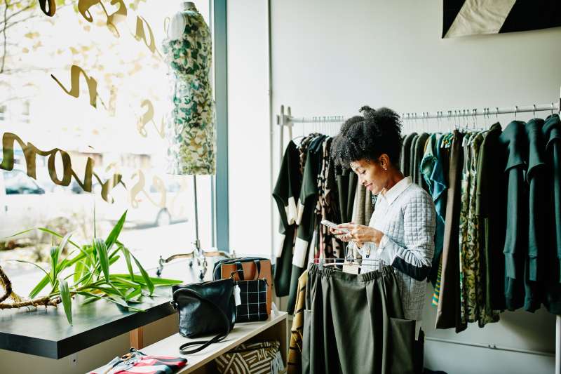 Woman o=inside a clothing boutique checking her phone and holding clothes