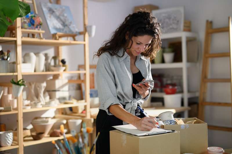 Mid adult potter woman, using smartphone and packing her products top boxes indoors in art studio, small business concept.