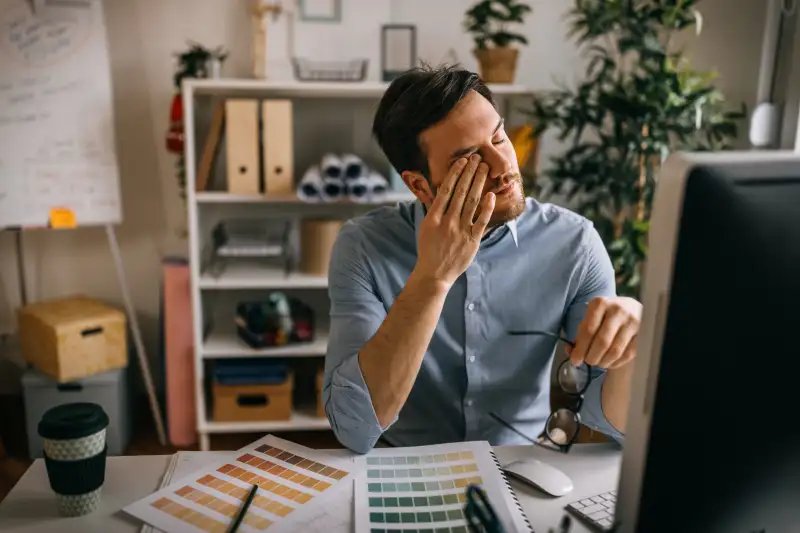 Exhausted male interior designer with color swatches and coffee cup sitting at desk in small office