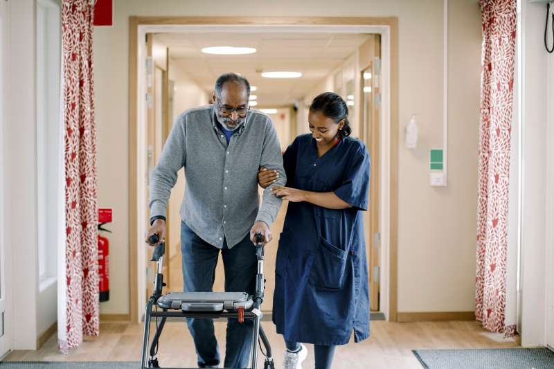 Smiling nurse holding hand of senior man walking with rollator in corridor at retirement home